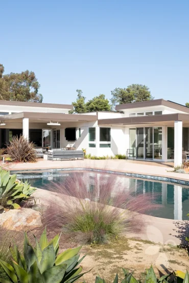 Poolside with white house and area with lounge chairs in background with bushes in front of the pool