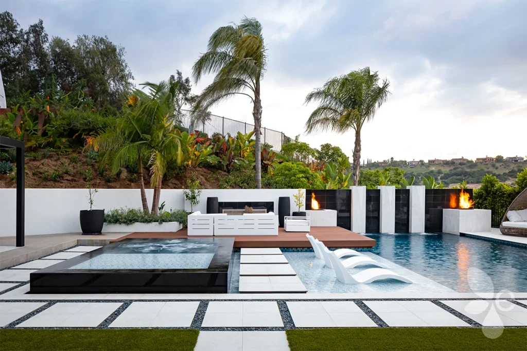 A backyard with a pool, water and fire features surrounded by Hardscape and Tropical Landscape