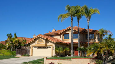 Mediterranean-style home featuring a red tile roof, stucco exterior, and lush landscaping with tall palm trees. The two-story house includes a spacious driveway and garage, reflecting a classic Southern California design in a residential neighborhood.